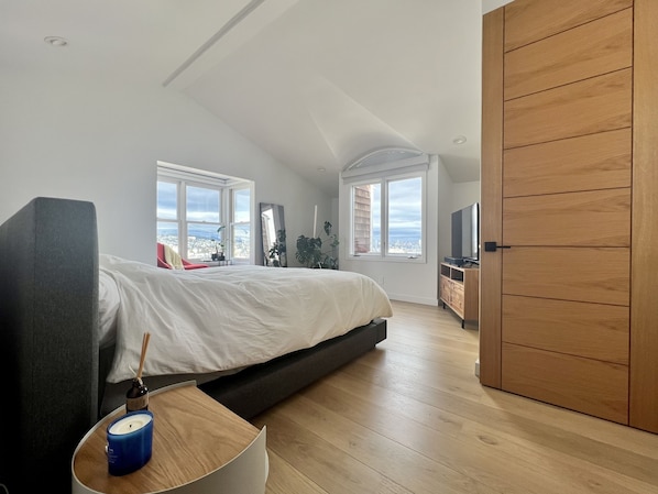 A modern bedroom with a large bed, a TV, and a city view through the windows.