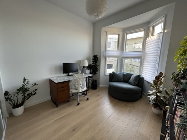 A living room with a wooden floor, a desk with a computer, a grey sofa, and a large window with blinds. - Live/Work House w/Views, Yard, Parking 3br/2ba (San Francisco)
