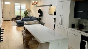 A modern kitchen with white cabinetry, a central island with stools, a dining area with a sofa, and a large window providing natural light. - Live/Work House w/Views, Yard, Parking 3br/2ba (San Francisco)