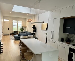 Una cuina moderna amb armaris blancs, una illa central amb tamborets, una zona de menjador amb sofà i un gran finestral que aporta llum natural.