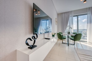 A modern living room with a large flat-screen TV, a dining area with green chairs, and a view of the city through large windows.