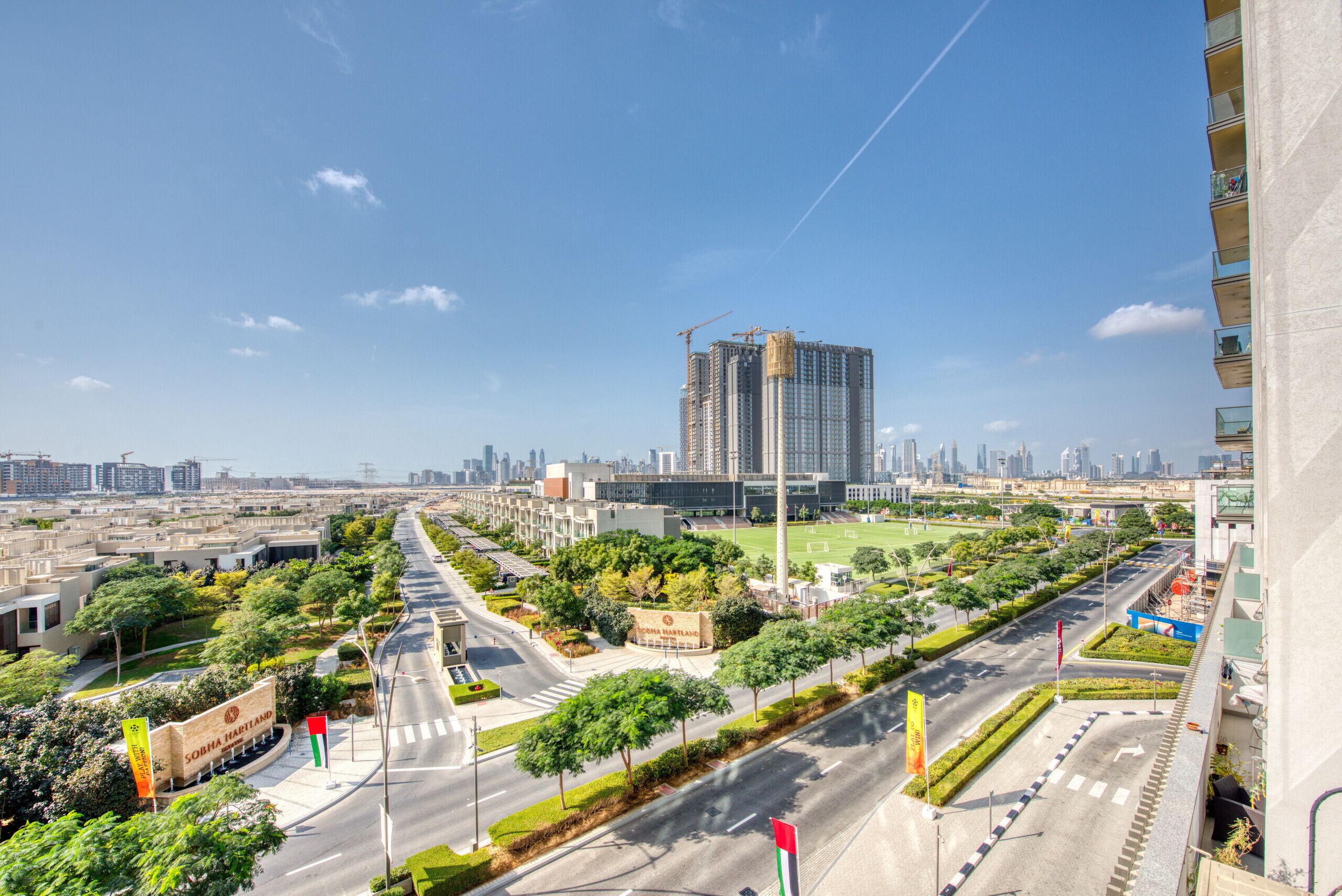 A modern urban landscape with a prominent high-rise building under construction, a well-maintained road with greenery, and a clear blue sky.