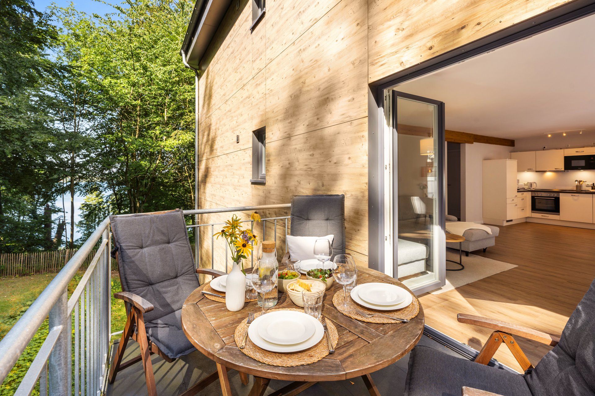 An outdoor dining area with a wooden table, chairs, and a glass door leading to a modern kitchen.