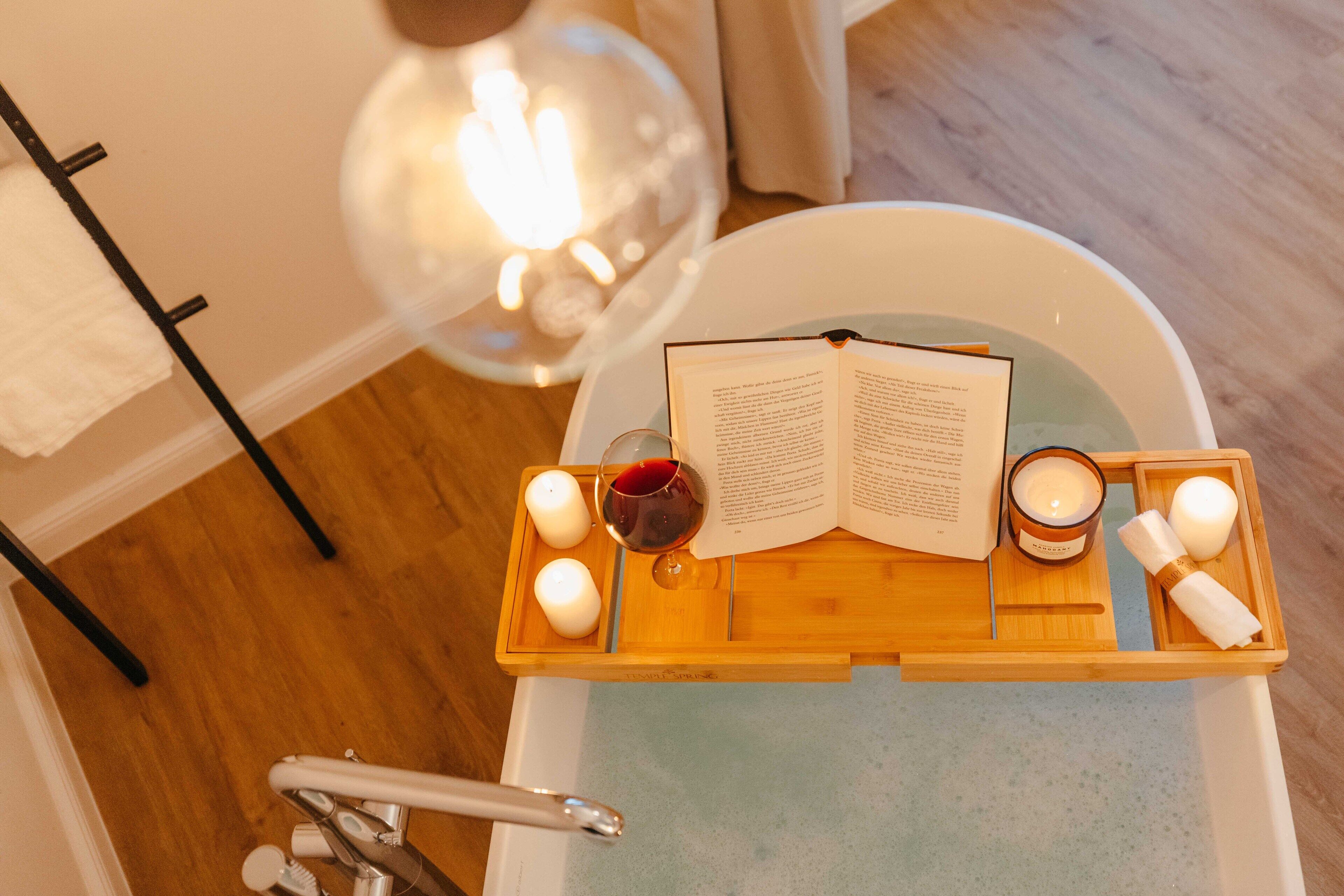 Ein hölzernes Tablett mit Kerzen, einem Buch und einem Glas Rotwein auf einer Badewanne.