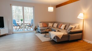Una sala de estar moderna con un sofá gris, una mesa de centro y un televisor sobre un mueble bajo. Hay un ventanal corredero que da a un área exterior con una mesa y sillas.