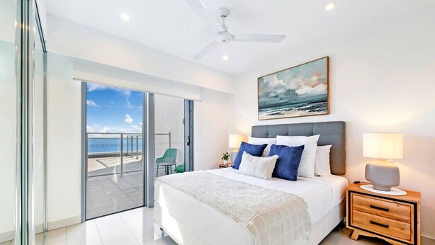A modern bedroom with a large bed, a ceiling fan, a painting of the sea, and a view of the ocean through a sliding door. - Zen Sea View 2BR, 2BA Penthouse (Darwin City)
