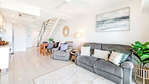 A modern living room with a grey sofa, a dining area with a staircase, and a large abstract painting on the wall. - Zen Sea View 2BR, 2BA Penthouse (Darwin City)