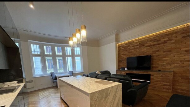 A modern kitchen with a marble island, wooden flooring, and a built-in TV on a brick wall.