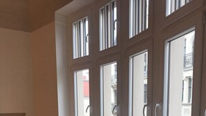 A room with a white table and two chairs, large windows with vertical blinds, and a view of buildings outside.