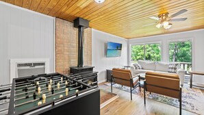 Una sala de estar acogedora con mesa de fútbol, chimenea, sofá cómodo y un gran ventanal con vistas a árboles.