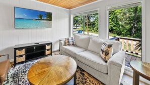 Una sala de estar con un televisor grande, un sofá, una mesa de centro y vistas a una playa tropical a través de grandes ventanales.