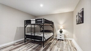 Cama litera en una habitación con suelo de madera, una mesita de noche pequeña con lámpara y un cuadro colgado en la pared.