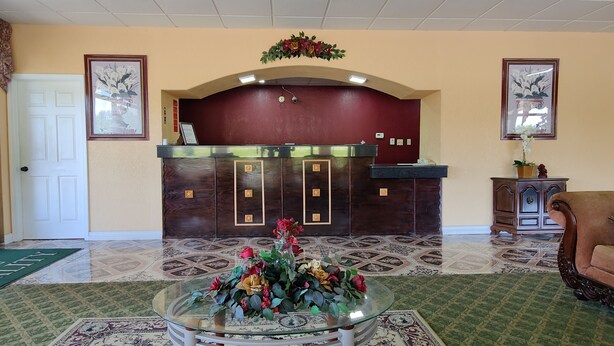 A reception area with a marble reception desk, a glass coffee table with flowers, patterned carpet, and a brown leather sofa. - Countryside Inn & Suites (Idabel)