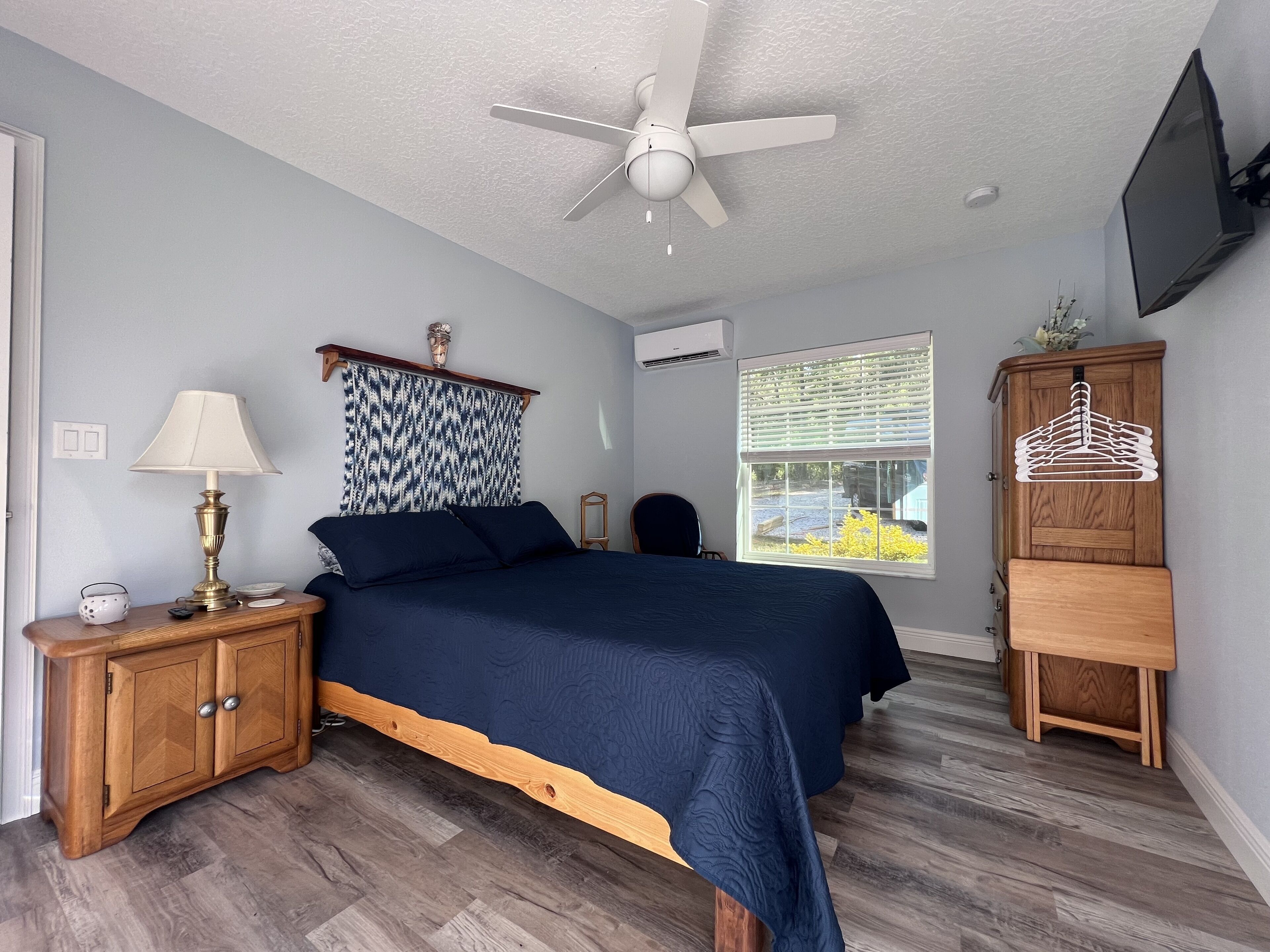 Une chambre à coucher comprenant un lit, un ventilateur de plafond, une fenêtre avec des stores, une armoire en bois et un téléviseur fixé au mur.