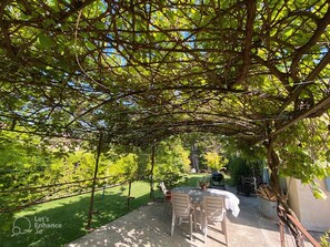 An outdoor patio with a table and chairs under a pergola, surrounded by lush greenery. - House with a Garden in Provence (Valréas)