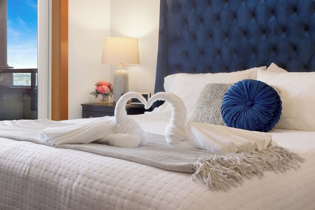 A neatly made bed with white linens, a blue tufted headboard, and a decorative pillow.