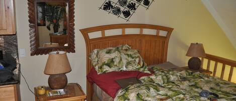 Un dormitorio con cabecera de madera, cama con edredón estampado con motivos tropicales, mesita de noche con lámpara, espejo y ventilador de techo.