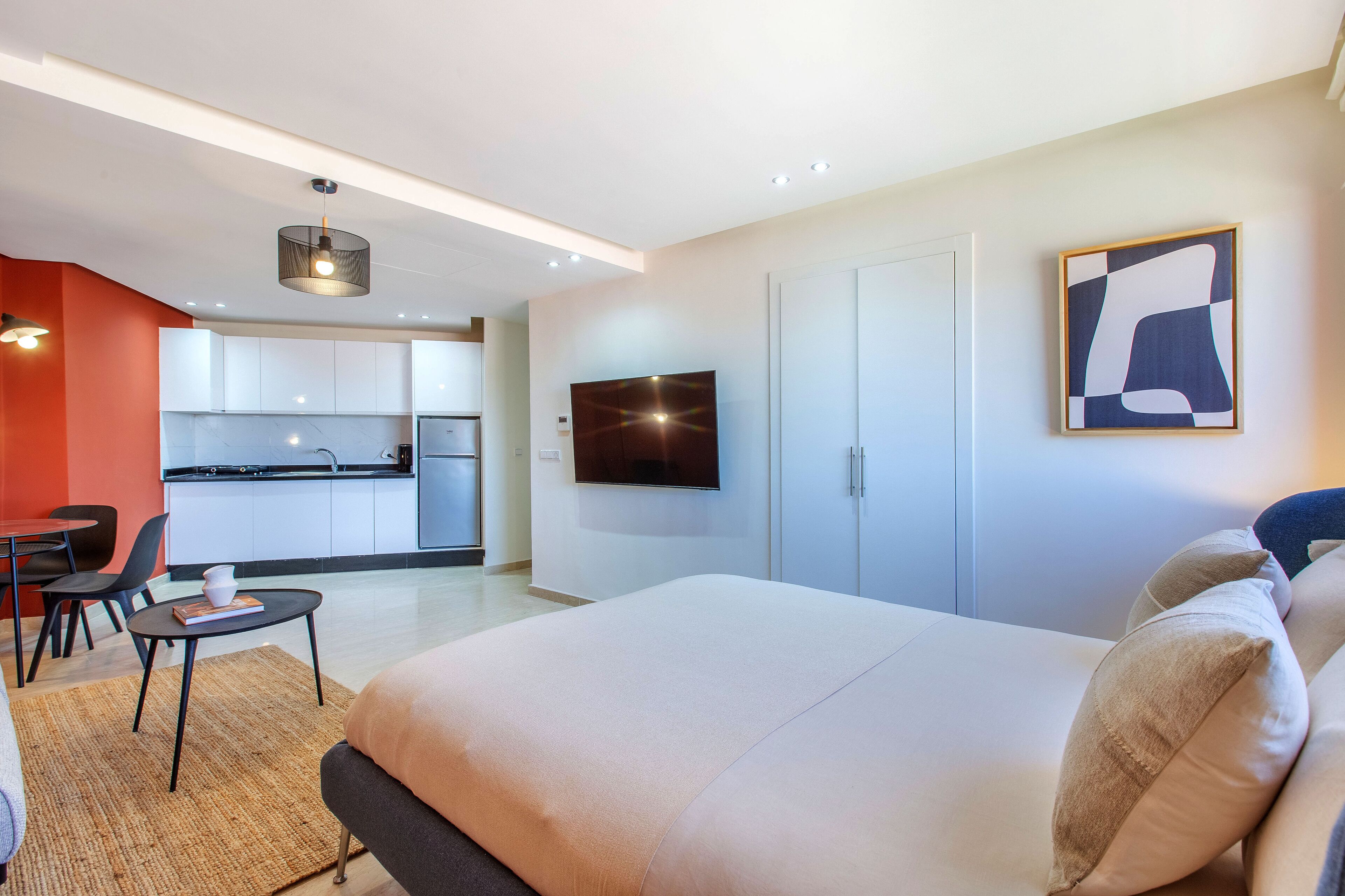 Una habitación de hotel moderna con una cama grande, un televisor de pantalla plana montado en la pared, una kitchenette al fondo y un área de comedor con una mesa y sillas.