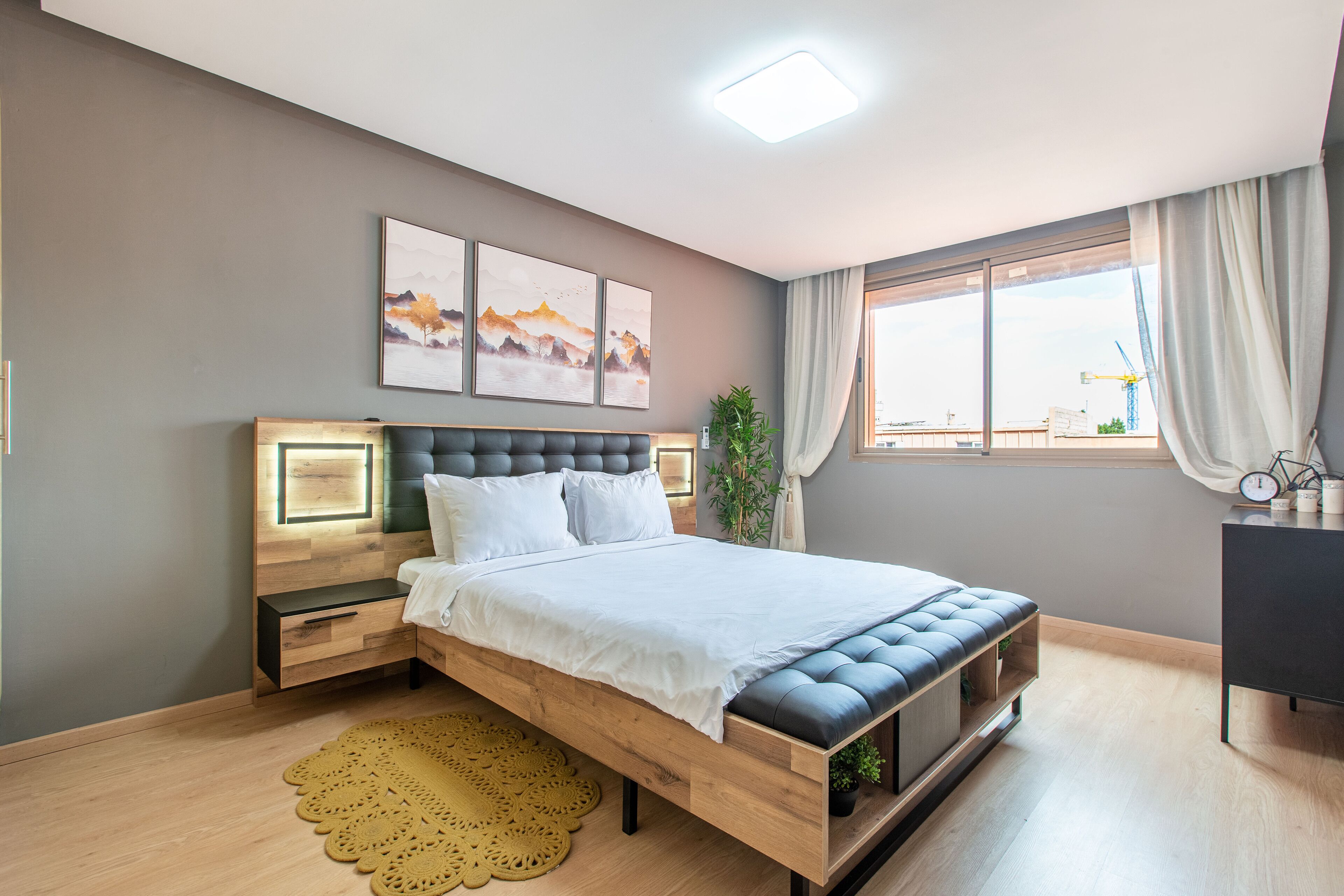 A modern bedroom with a wooden bed, bedside tables, a large window with curtains, and artwork on the wall.