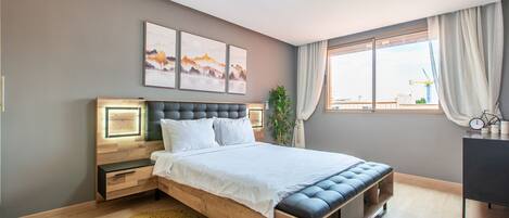 A modern bedroom with a wooden bed, bedside tables, a large window with curtains, and artwork on the wall.