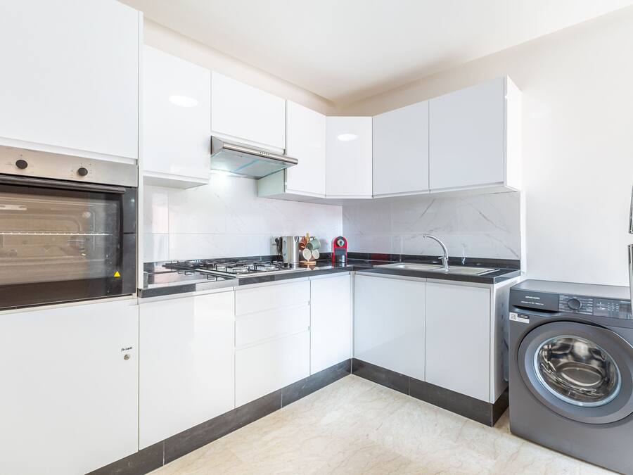Una cocina moderna con gabinetes blancos, horno empotrado, estufa a gas, lavamanos y lavadora de carga frontal.