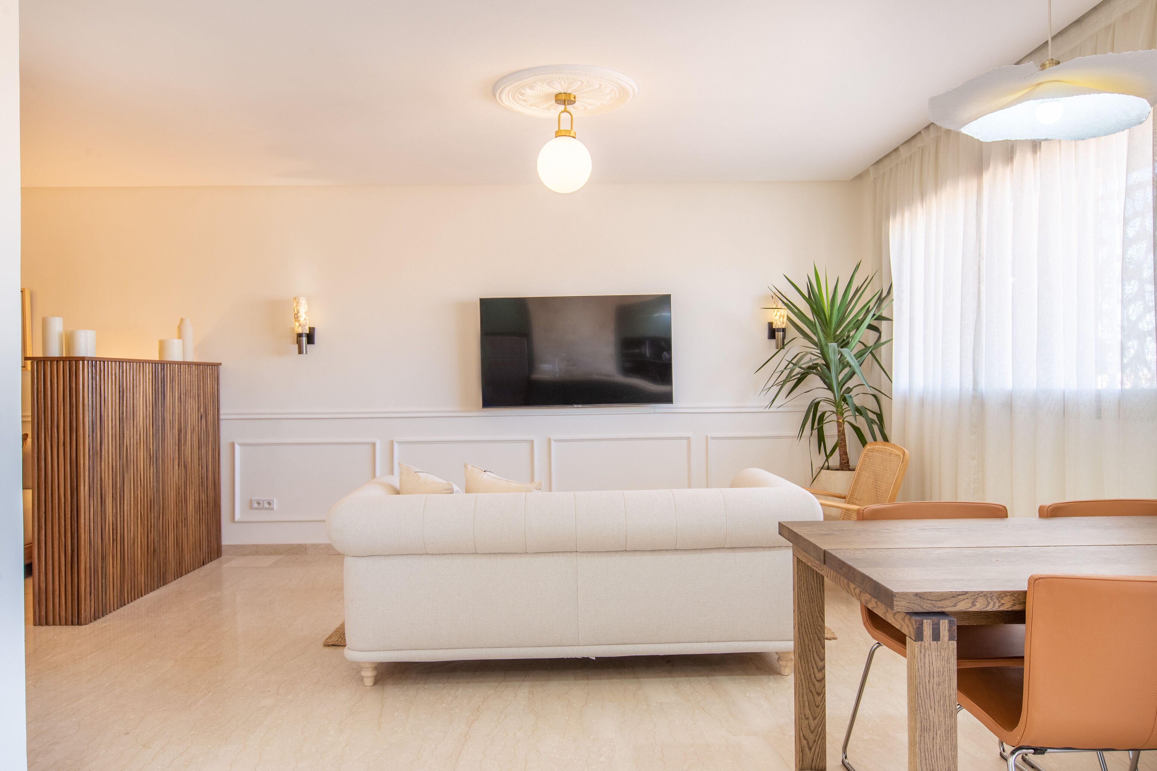 A modern living room with a flat-screen TV, a white sofa, a wooden dining table, and a potted plant.