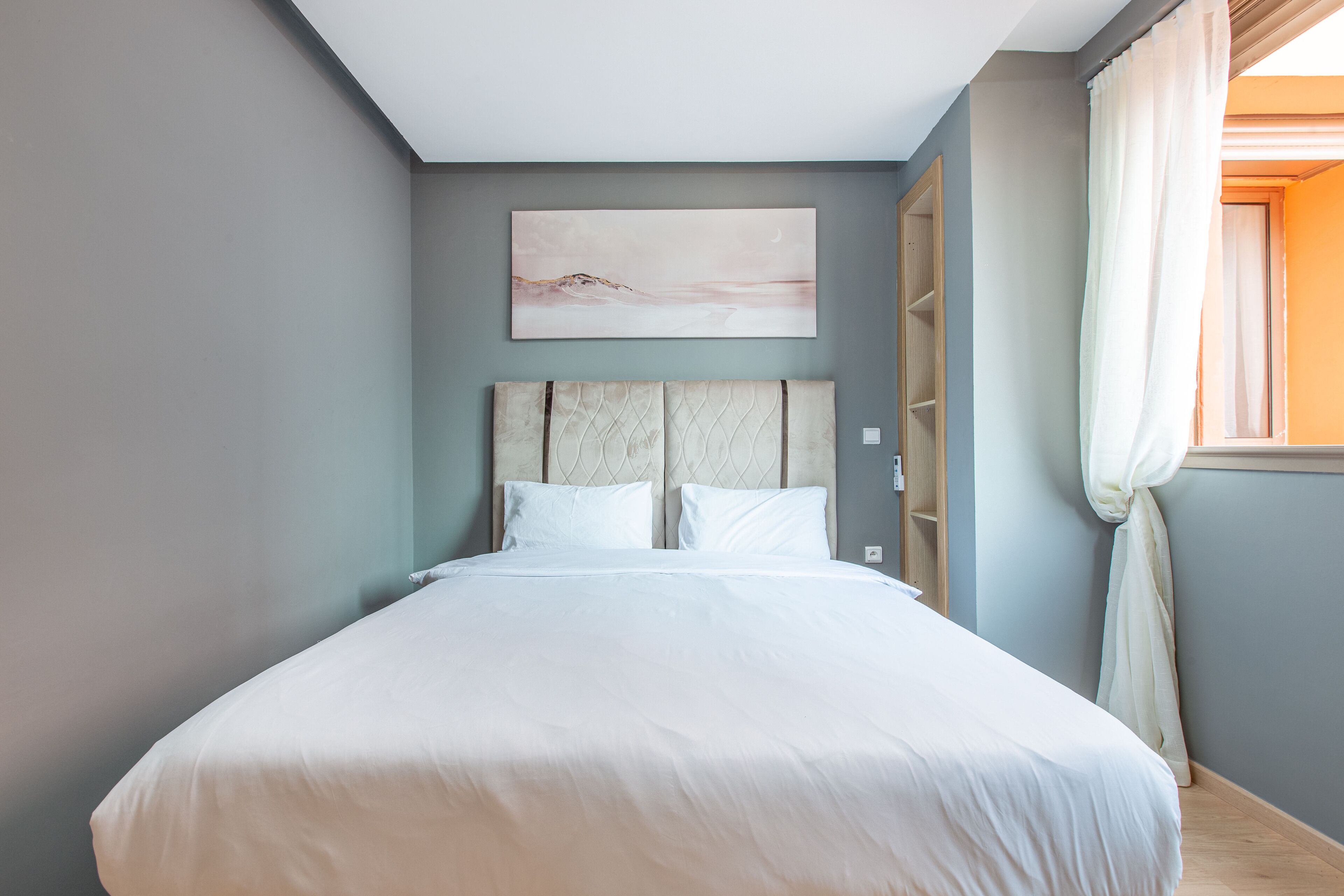 Une chambre à coucher avec un lit, une tête de lit, un tableau accroché au mur et une fenêtre avec des rideaux.