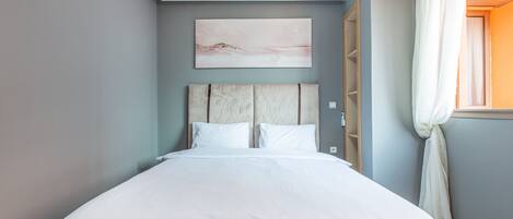 Une chambre à coucher avec un lit, une tête de lit, un tableau accroché au mur et une fenêtre avec des rideaux.