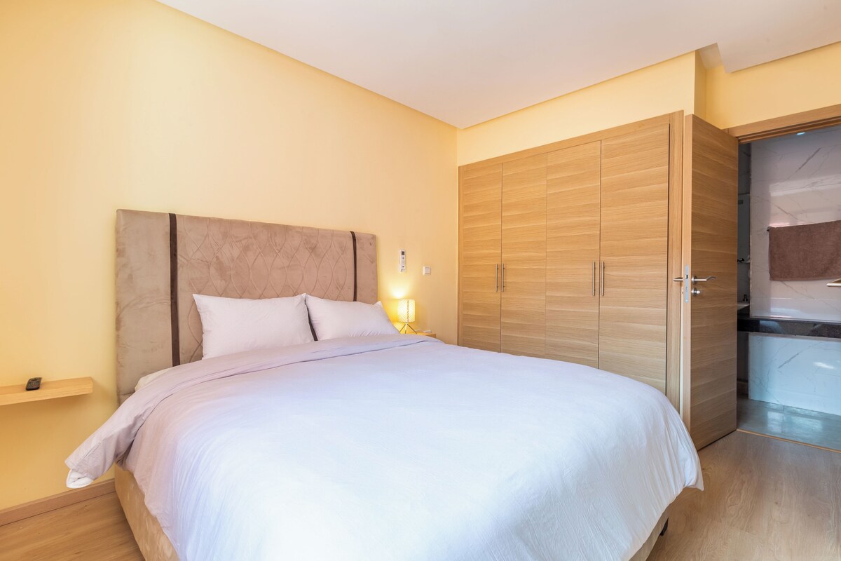 A bedroom with a bed, bedside table, lamp, wardrobe, and a bathroom visible through an open door.