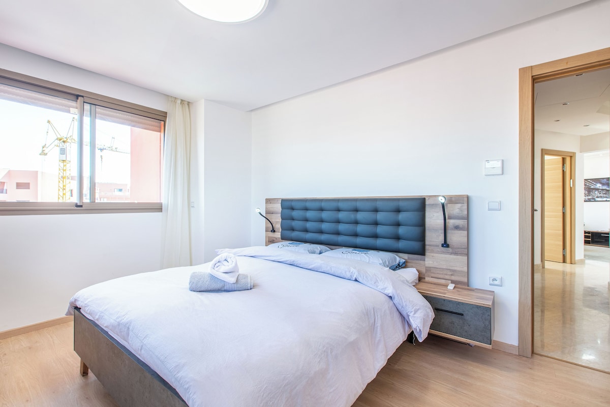 A modern bedroom with a bed, bedside tables, and a window with a view of buildings.