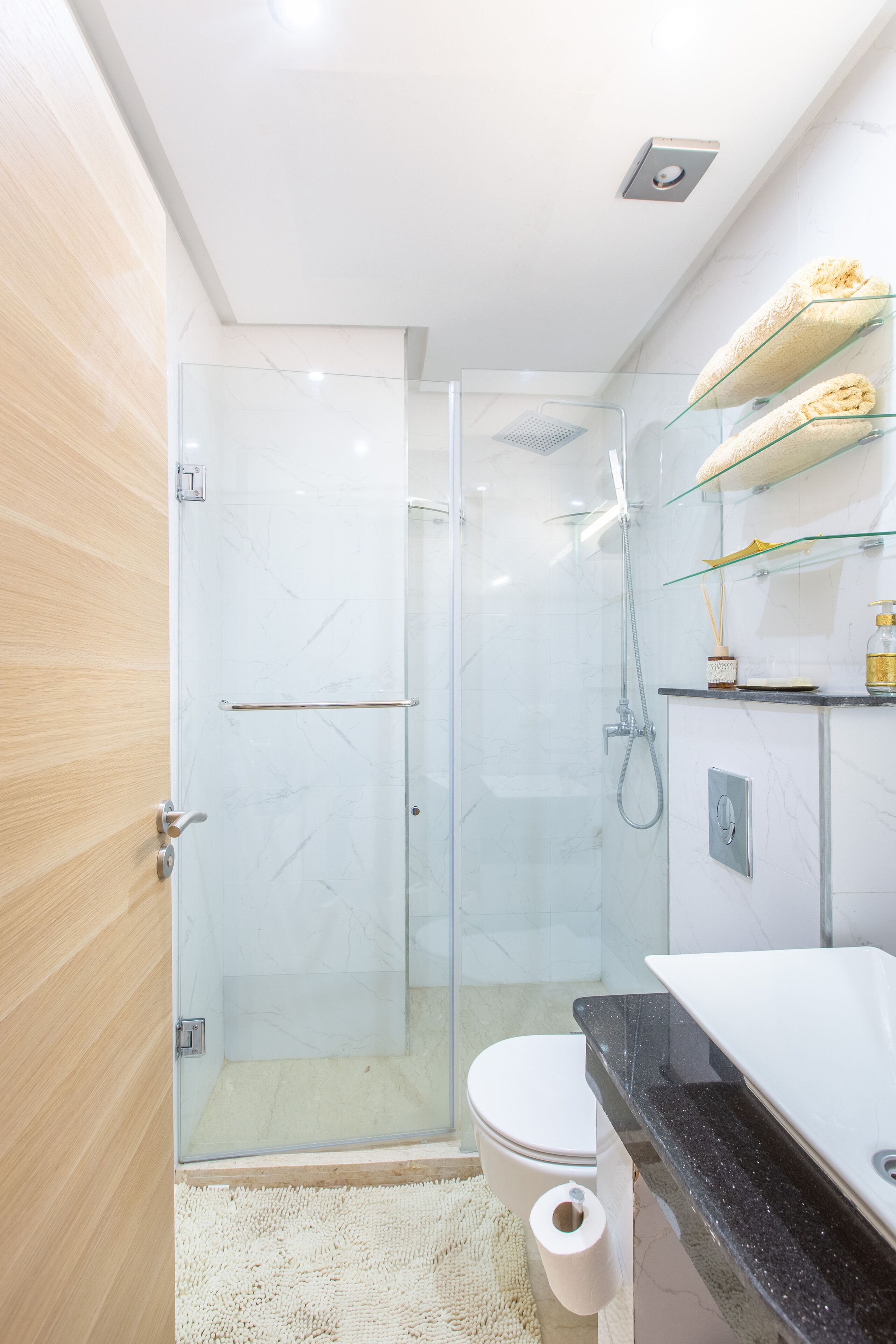 A modern bathroom with a glass shower enclosure, a toilet, a sink with a black countertop, and a white marble wall.