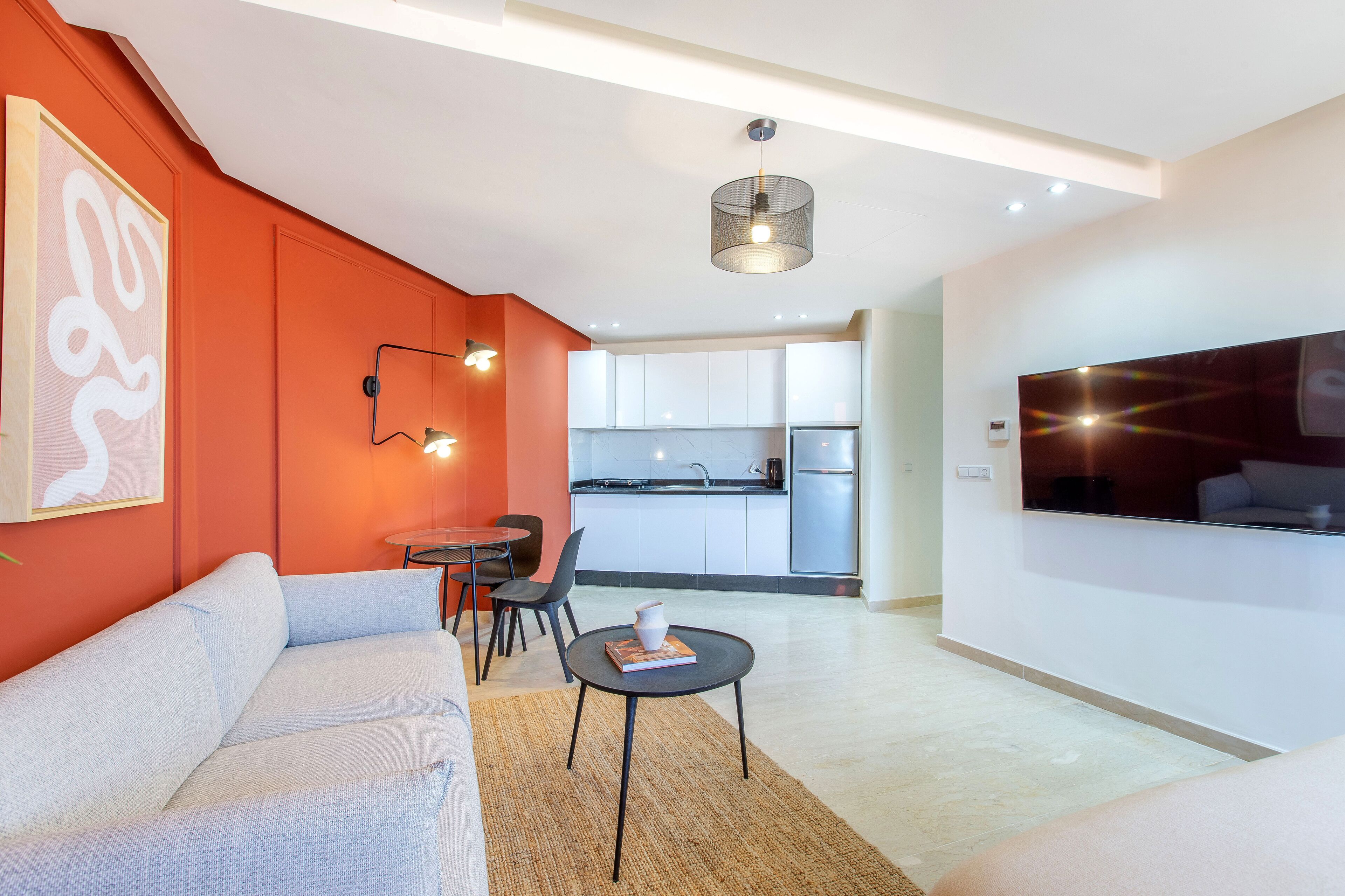 Una sala de estar moderna con un sofá, una mesa de centro y un televisor. Se aprecia una zona de cocina con un refrigerador y un lavabo.