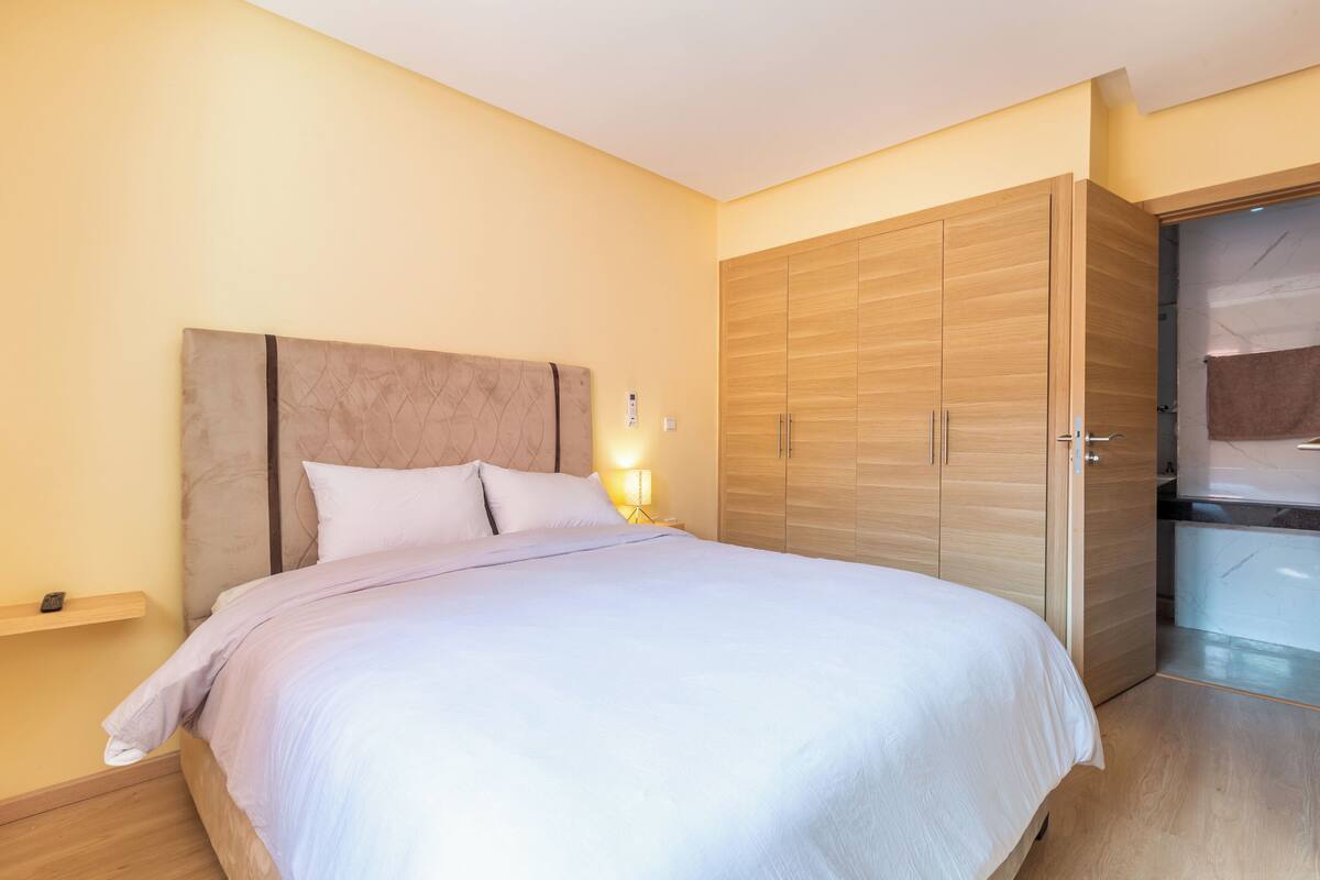 A modern bedroom with a large bed, a wooden wardrobe, and a bedside table with a lamp.