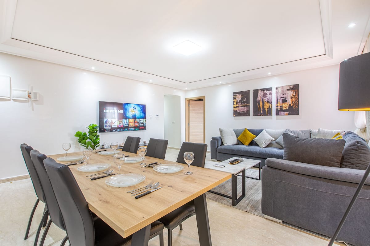 A modern living room with a dining table set for six, a grey sofa, and a flat-screen TV mounted on the wall.