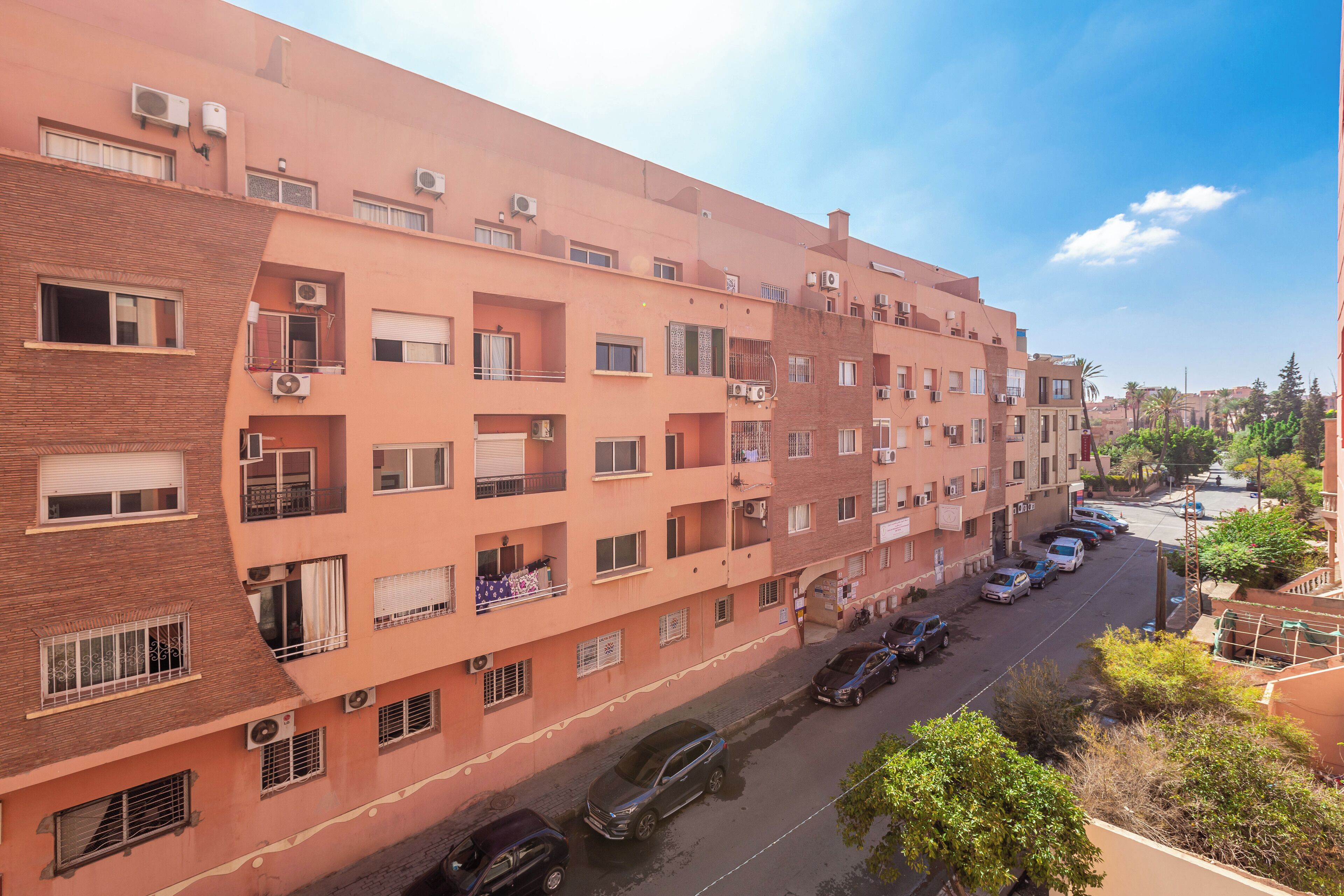 A multi-story residential building with balconies, air conditioning units, and a parking area with cars.