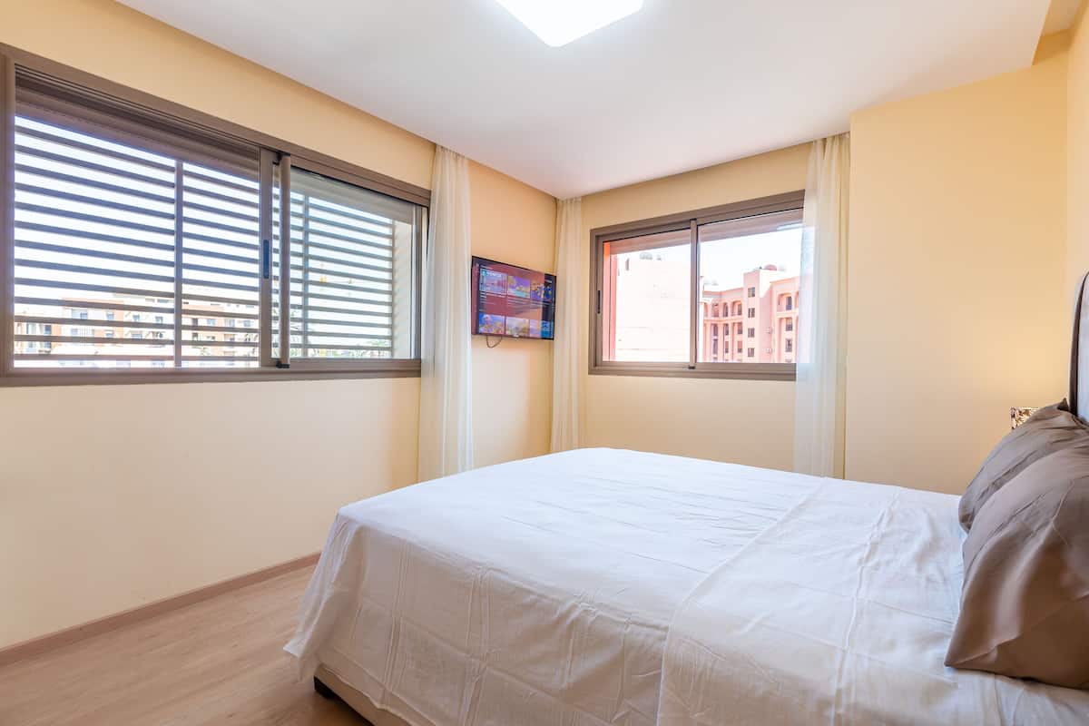 A bedroom with a bed, two windows, and a TV on the wall.
