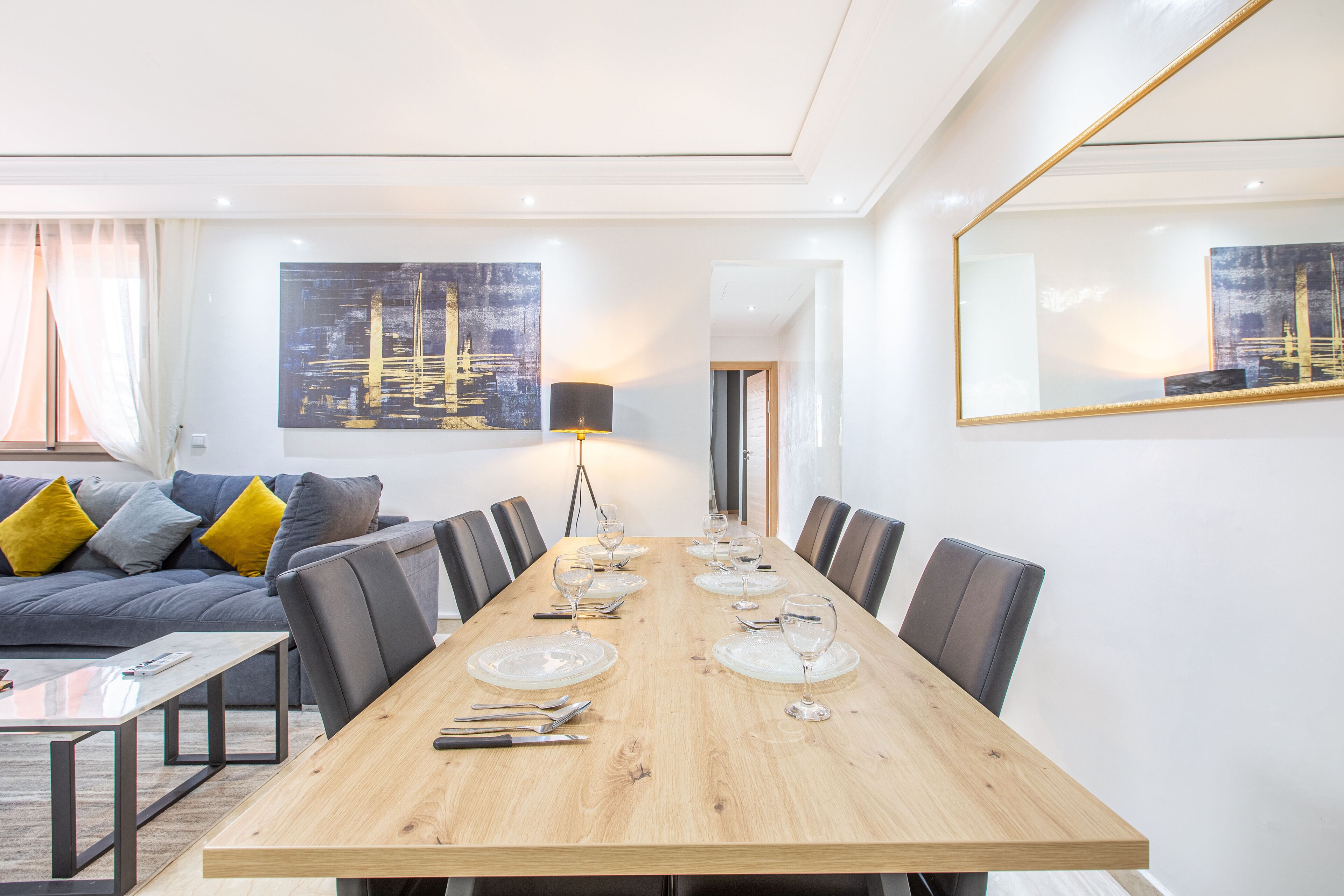 A modern dining area with a long wooden table set for six, surrounded by chairs, in a well-lit room with a sofa and cushions in the background.