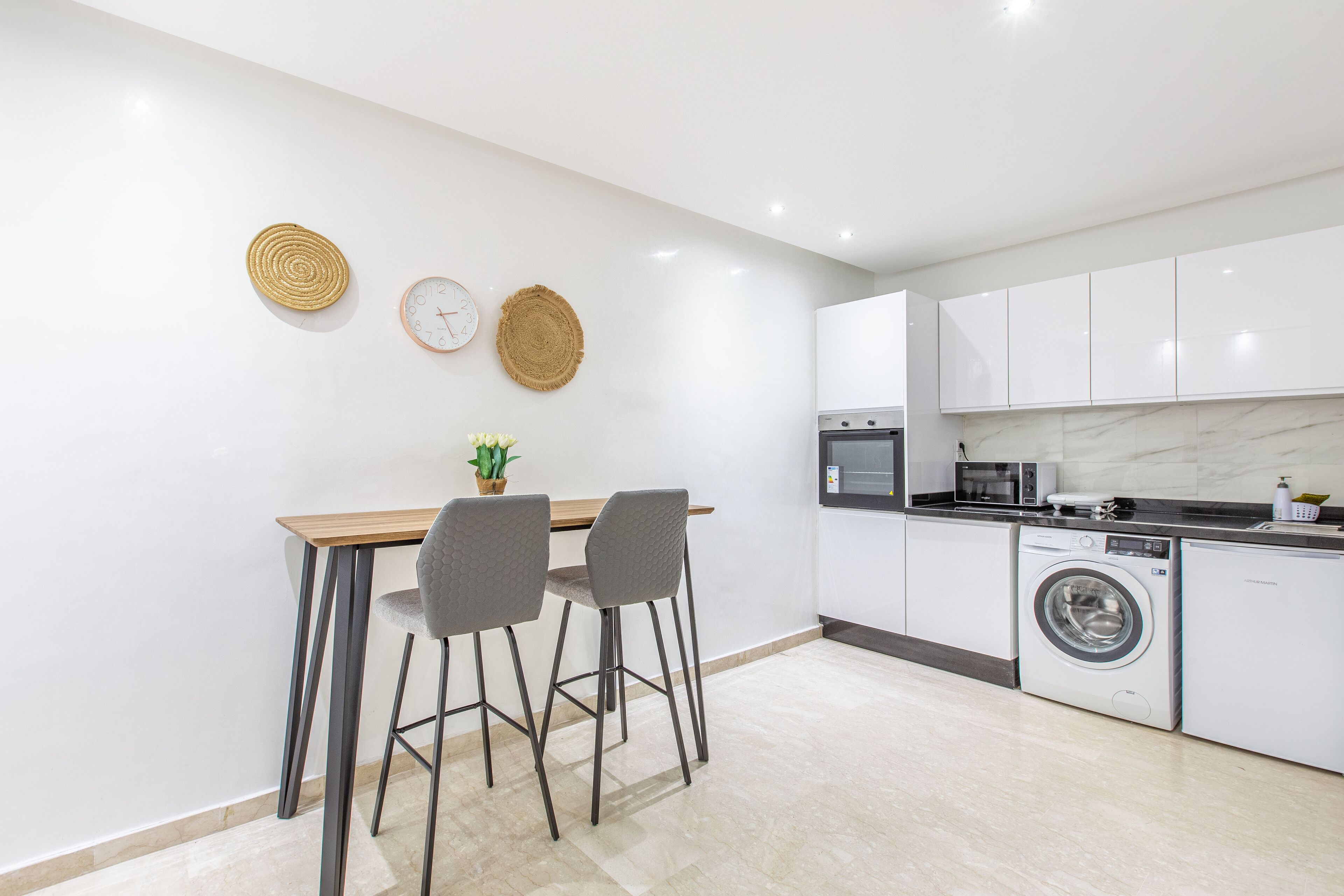 A modern kitchen with a dining area, featuring a microwave, oven, washing machine, and a clock on the wall.