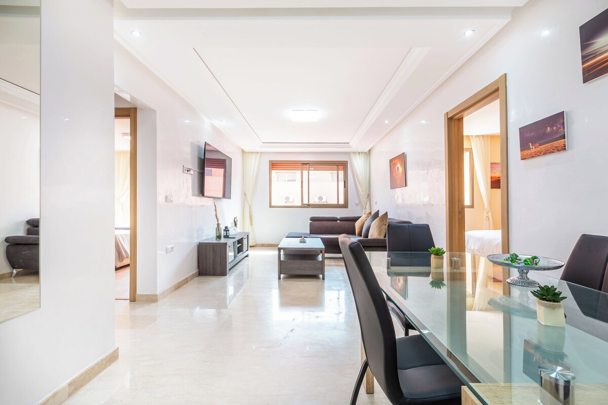 A modern living room with a glass dining table, a sofa, and a TV mounted on the wall.