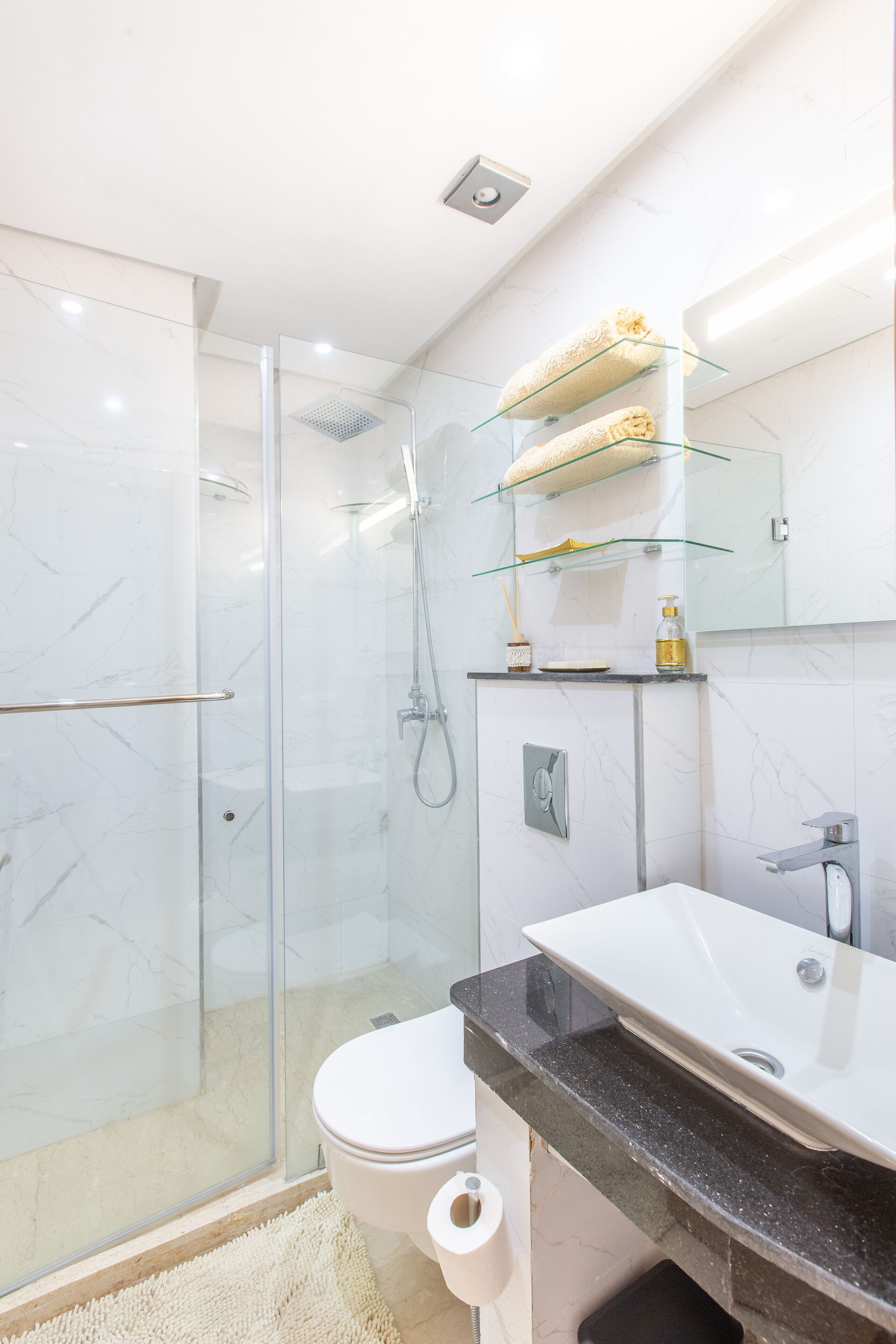 Une salle de bain moderne avec une douche à l’italienne en verre, des toilettes blanches, un lavabo noir et un plan de toilette en verre où sont posés les articles de toilette.