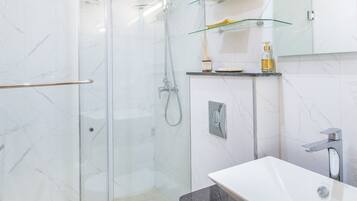 Une salle de bain moderne avec une douche à l’italienne en verre, des toilettes blanches, un lavabo noir et un plan de toilette en verre où sont posés les articles de toilette.