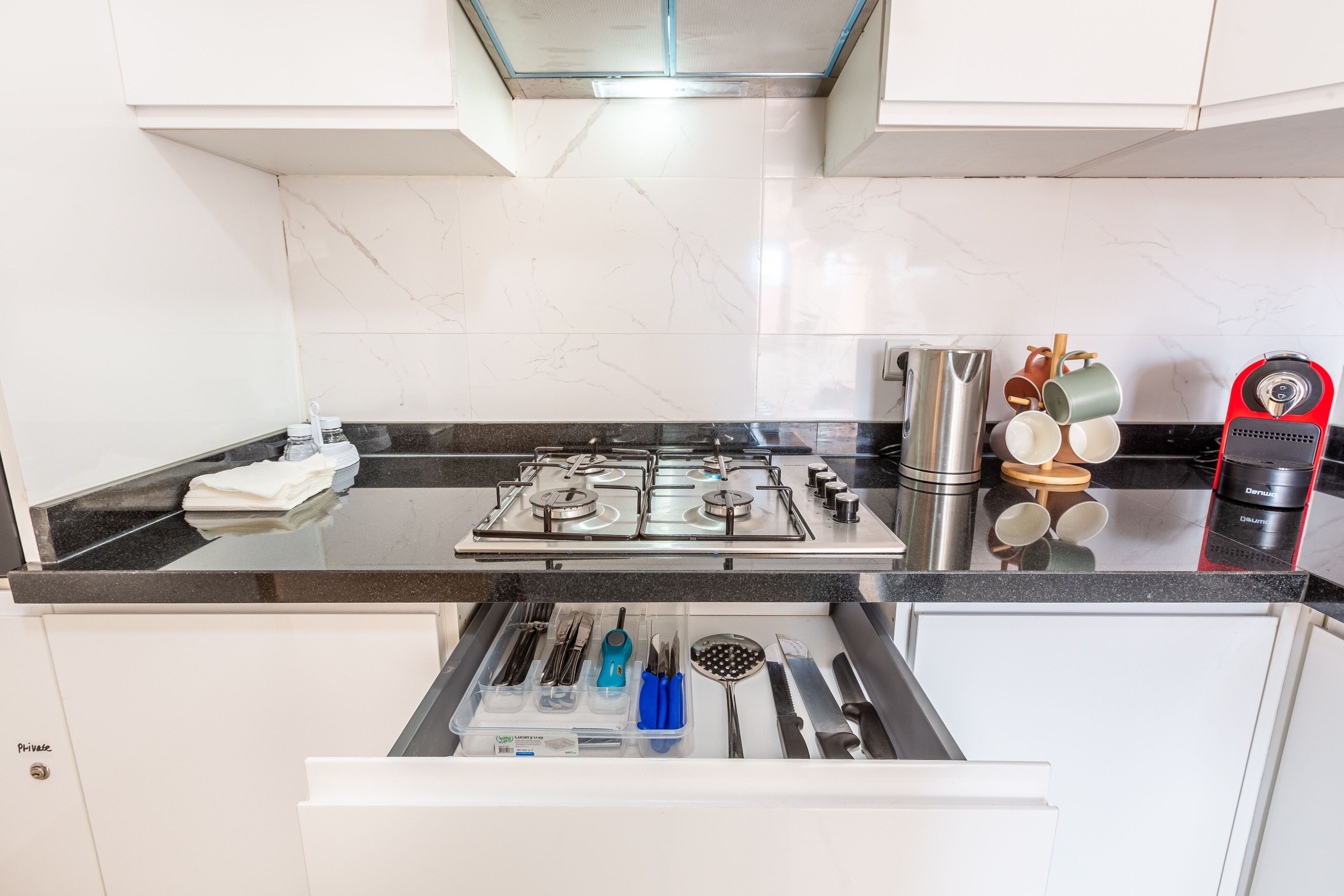 A kitchen with a gas stove, a coffee maker, and a drawer with utensils.