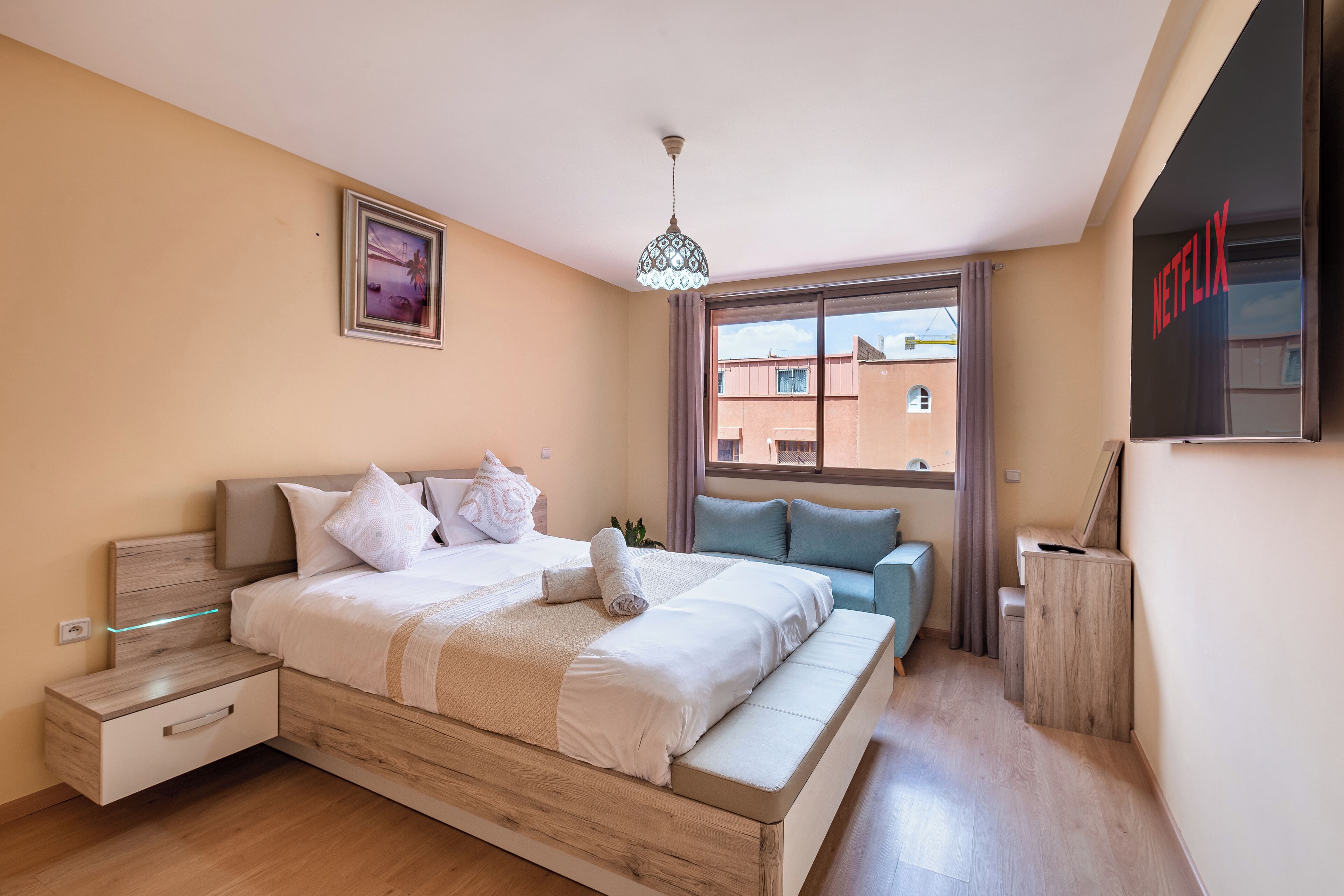A modern bedroom with a wooden bed, a bedside table, a wall-mounted TV, and a window with a view of buildings outside.