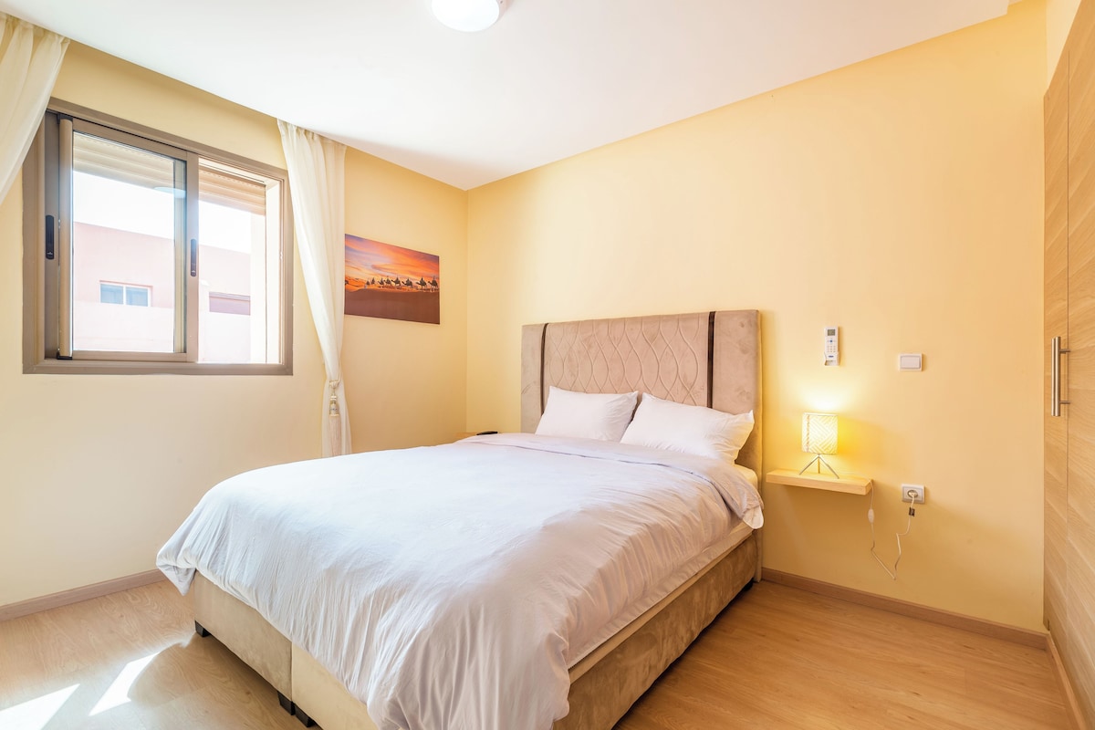 A hotel room with a bed, a nightstand, a lamp, a painting, and a window with curtains.
