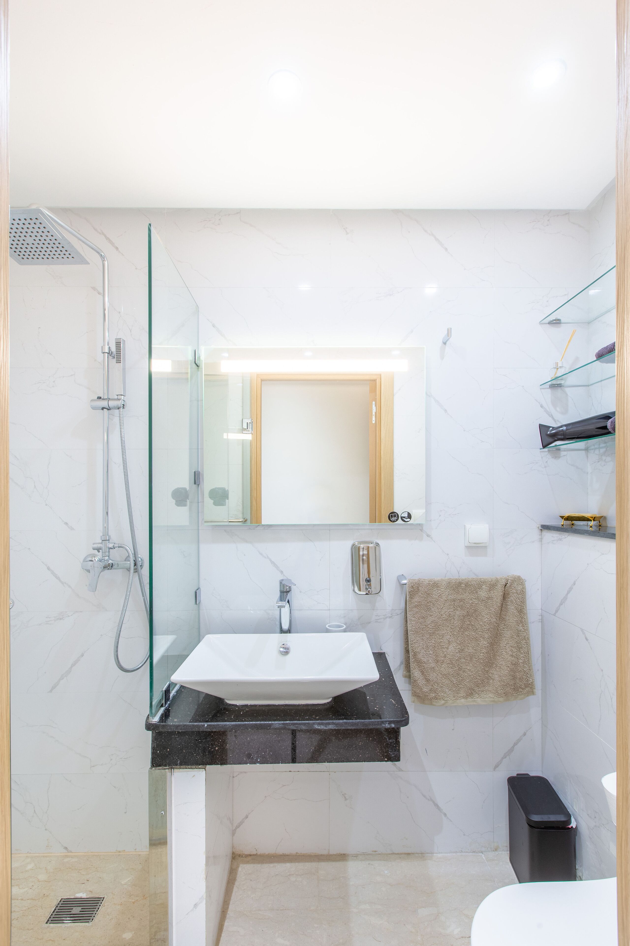 Un baño moderno con cabina de ducha de vidrio, un lavabo blanco con encimera negra, un espejo sobre el lavabo y una toalla colgada en la pared.