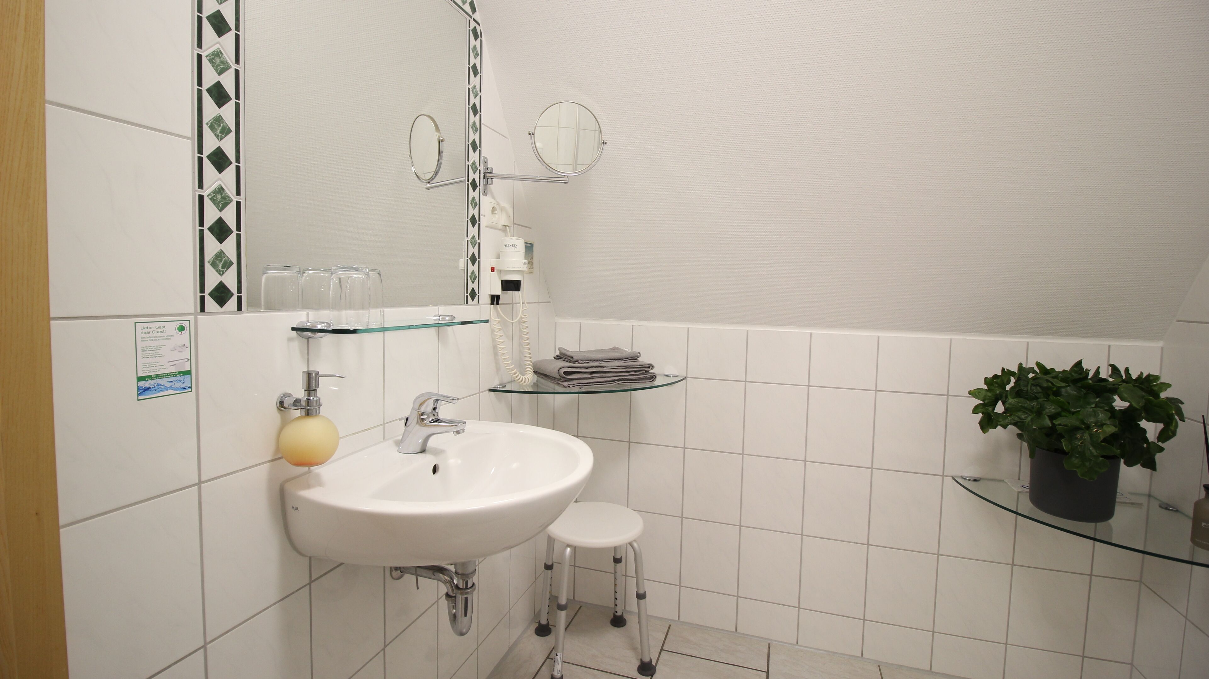 Un baño con un lavamanos blanco, un estante de vidrio, un taburete pequeño, un espejo y una planta en maceta.