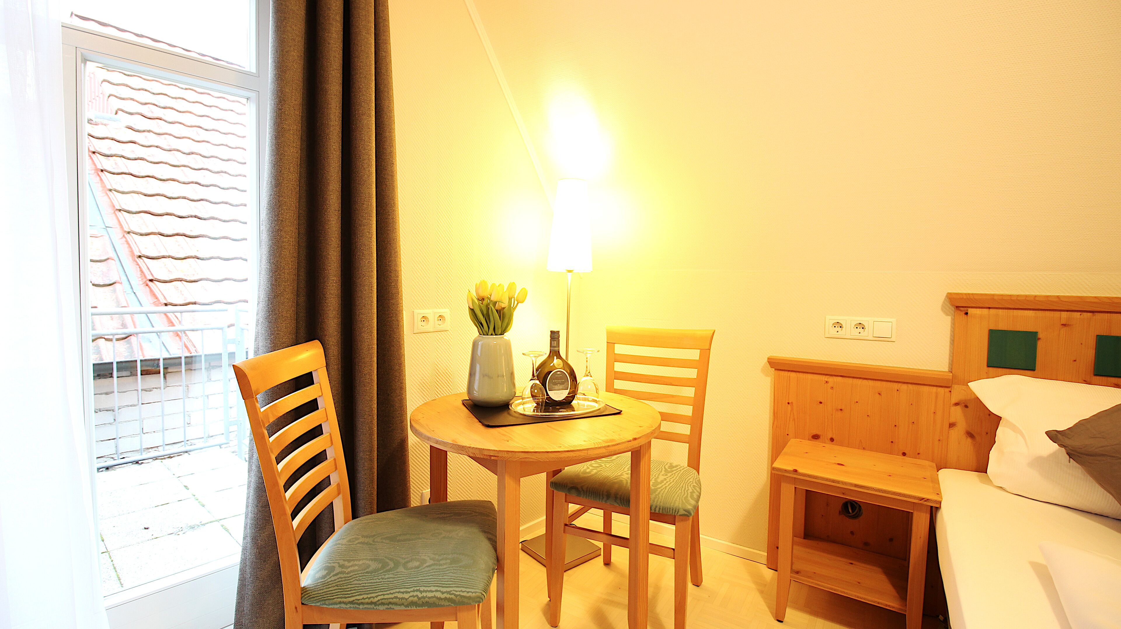 A hotel room with a wooden table, two chairs, a lamp, a bottle, and a vase with flowers.