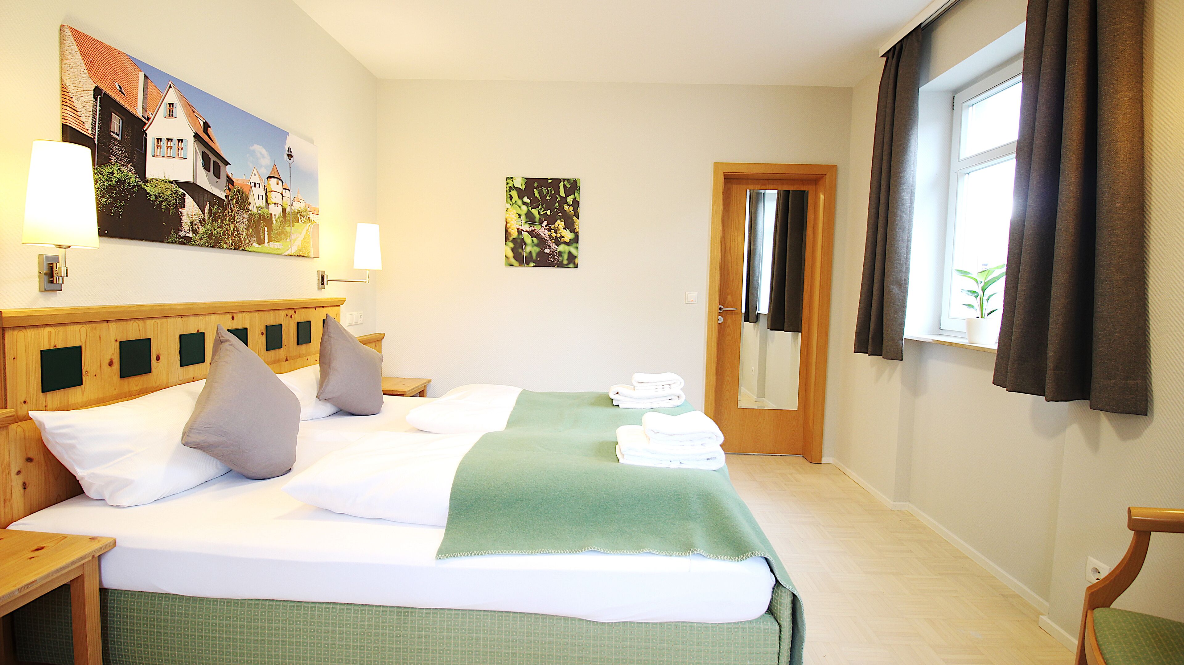 A hotel room with a bed, pillows, a green bedspread, a wooden headboard, a nightstand, a wall-mounted lamp, a painting, a door, and a window with curtains.