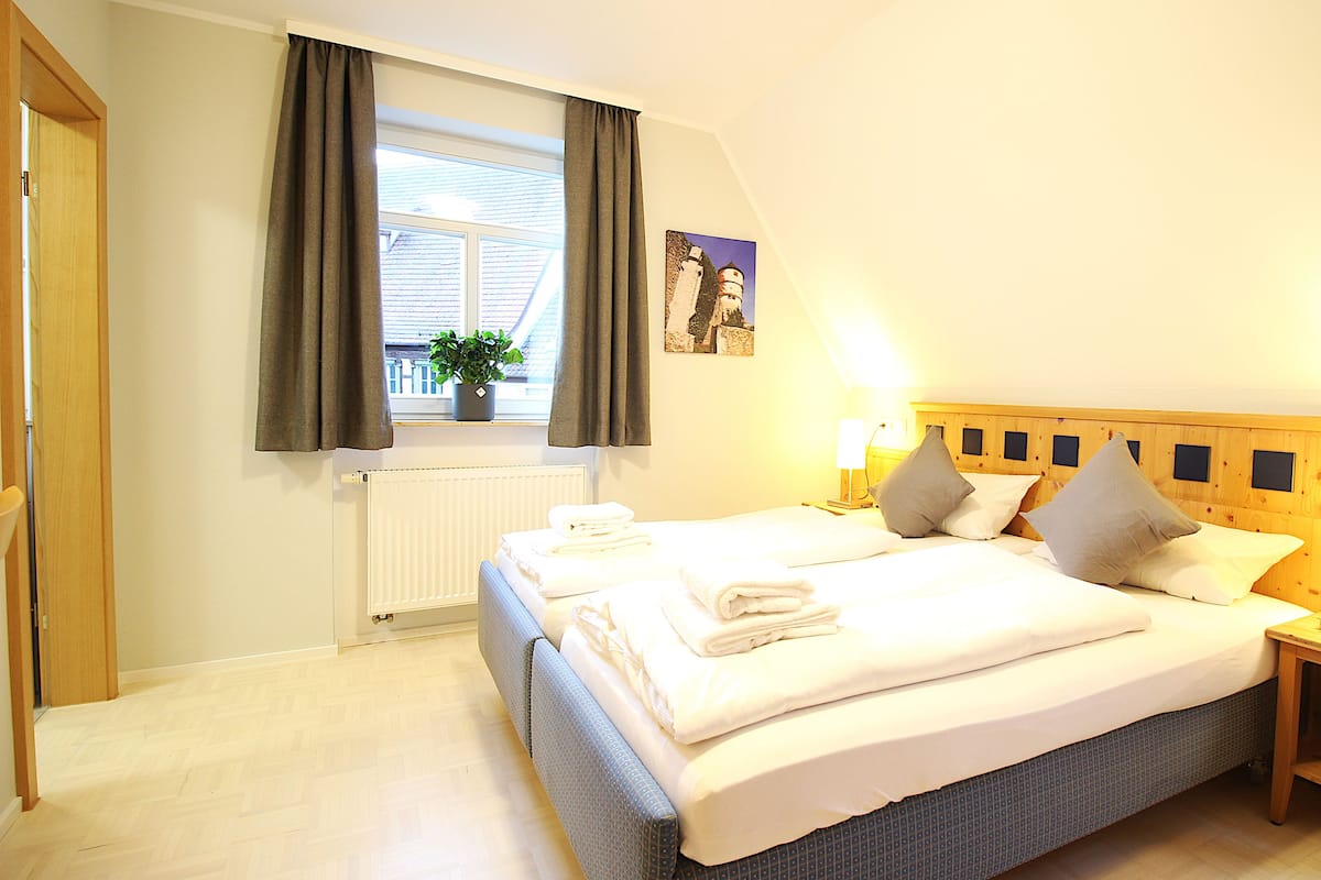 A hotel room with a large bed, two bedside tables with lamps, a window with curtains, a radiator, and a potted plant on the windowsill.