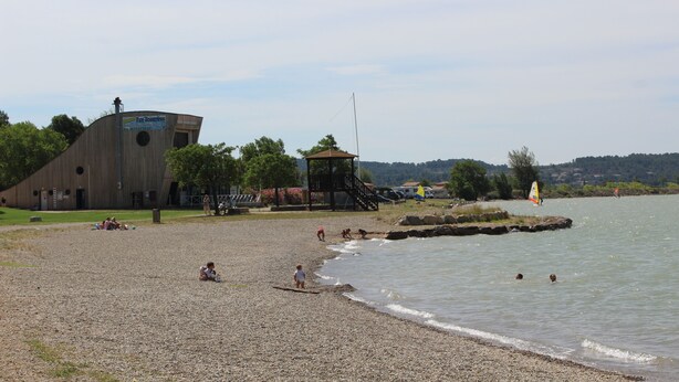 Ein Kiesstrand mit einem Holzgebäude, einem Rettungsturm und einem Segelboot im Wasser.